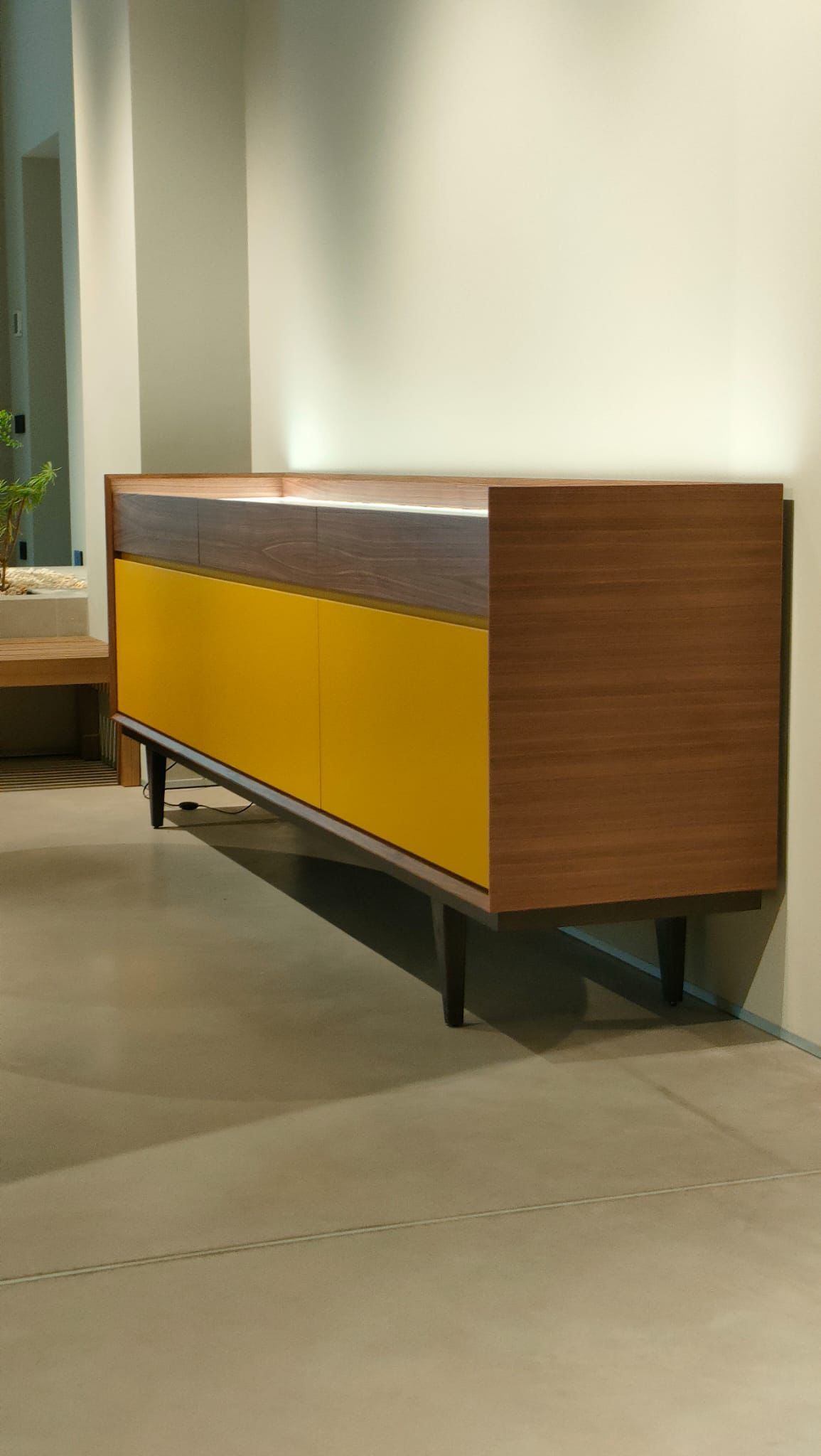 Una credenza in stile moderno di metà secolo con struttura effetto legno, ante gialle e gambe scure, poggiata su un pavimento piastrellato.