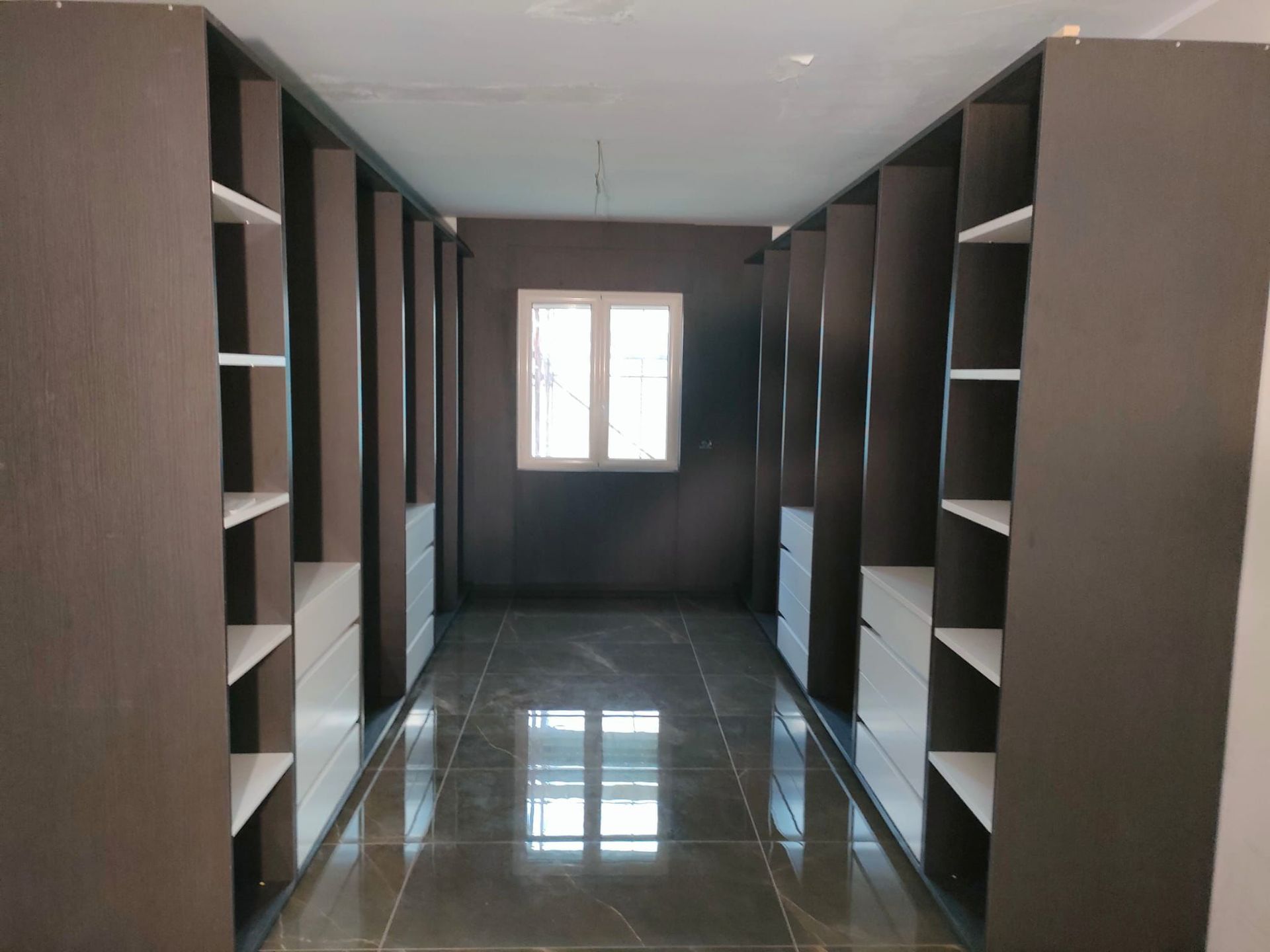 Una cabina armadio con ripiani in legno scuro, cassetti bianchi e un pavimento piastrellato lucido che conduce a una finestra.