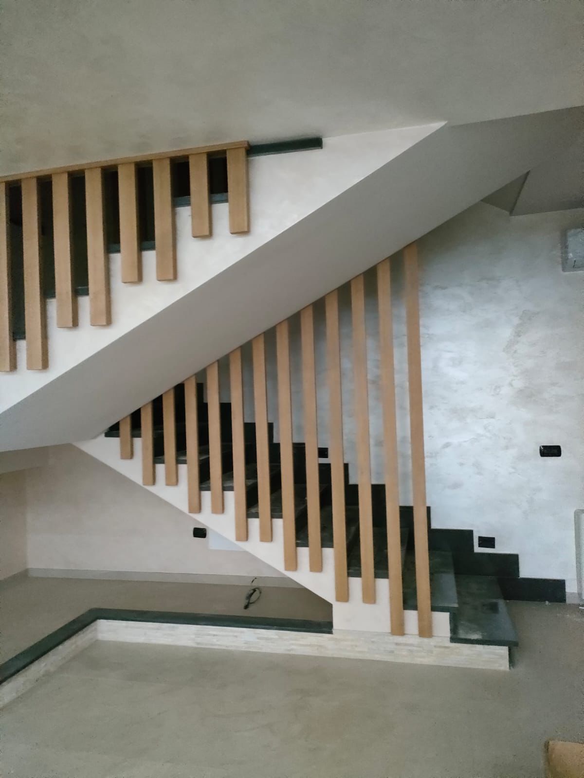 Una scala moderna con gradini in pietra scura e listelli verticali in legno che fungono da ringhiera, su una parete bianca con finitura materica.