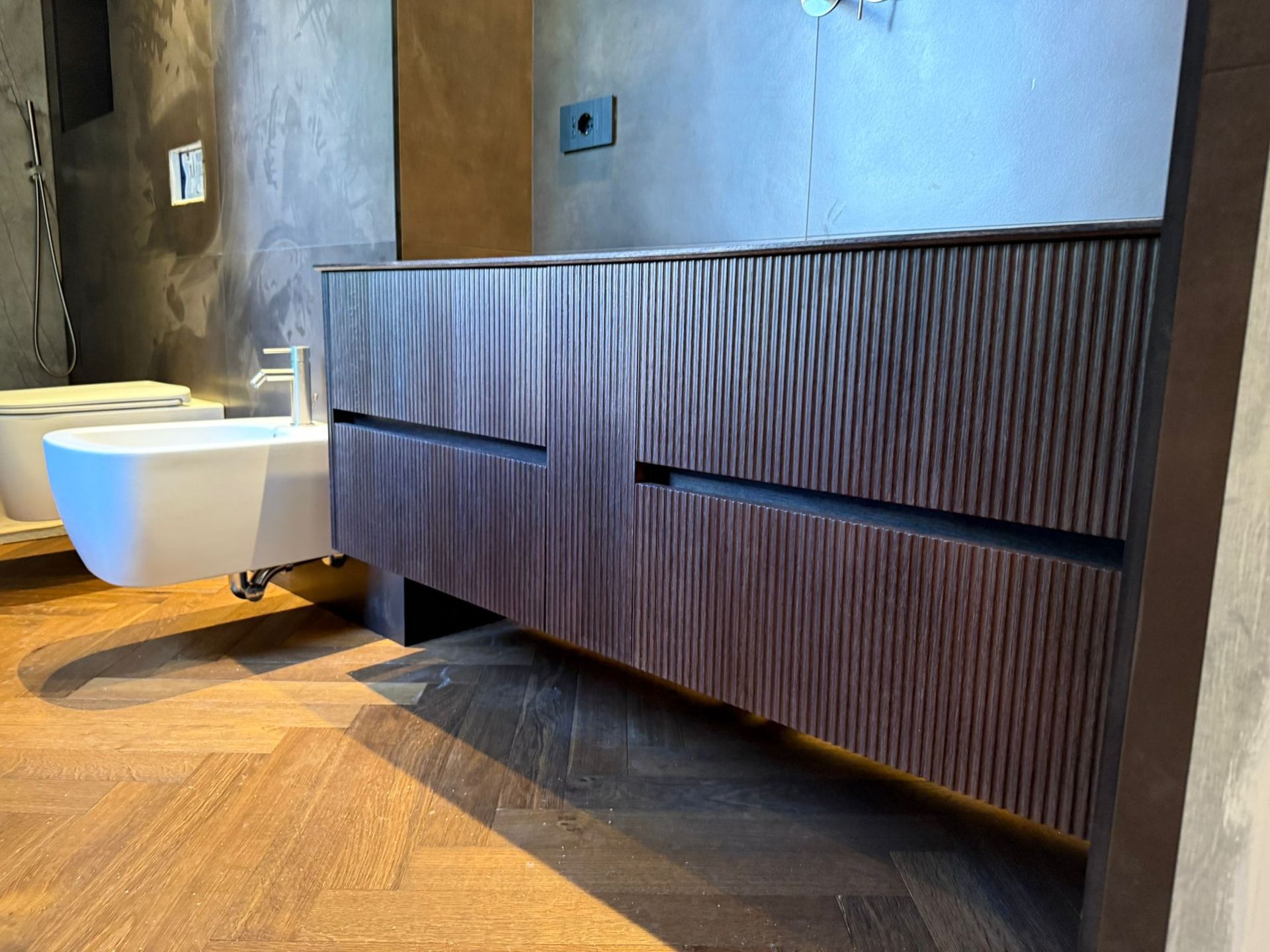 Un mobiletto da bagno in legno scuro a coste, montato a parete sopra un pavimento in legno scuro a spina di pesce.