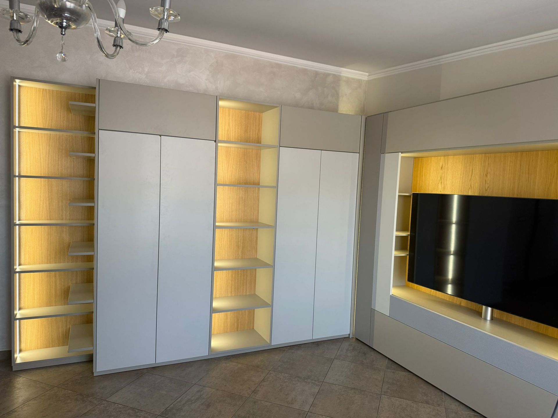 Mobili su misura moderni con ripiani a giorno illuminati e ante bianche chiuse, accanto a un televisore montato a parete.
