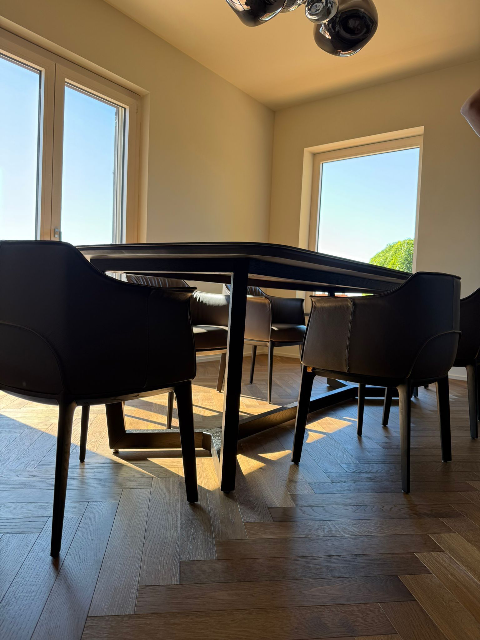 Un tavolo da pranzo scuro circondato da poltrone imbottite scure in una stanza luminosa con pavimento in legno e ampie finestre.