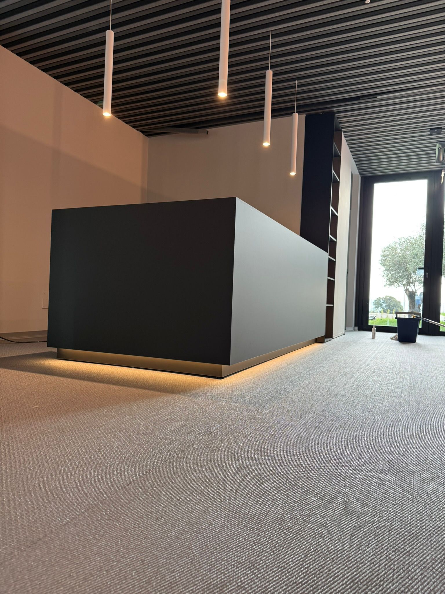 In una stanza moderna illuminata da lampade a sospensione, un bancone reception minimalista di colore grigio scuro poggia su un pavimento ruvido e decorato.