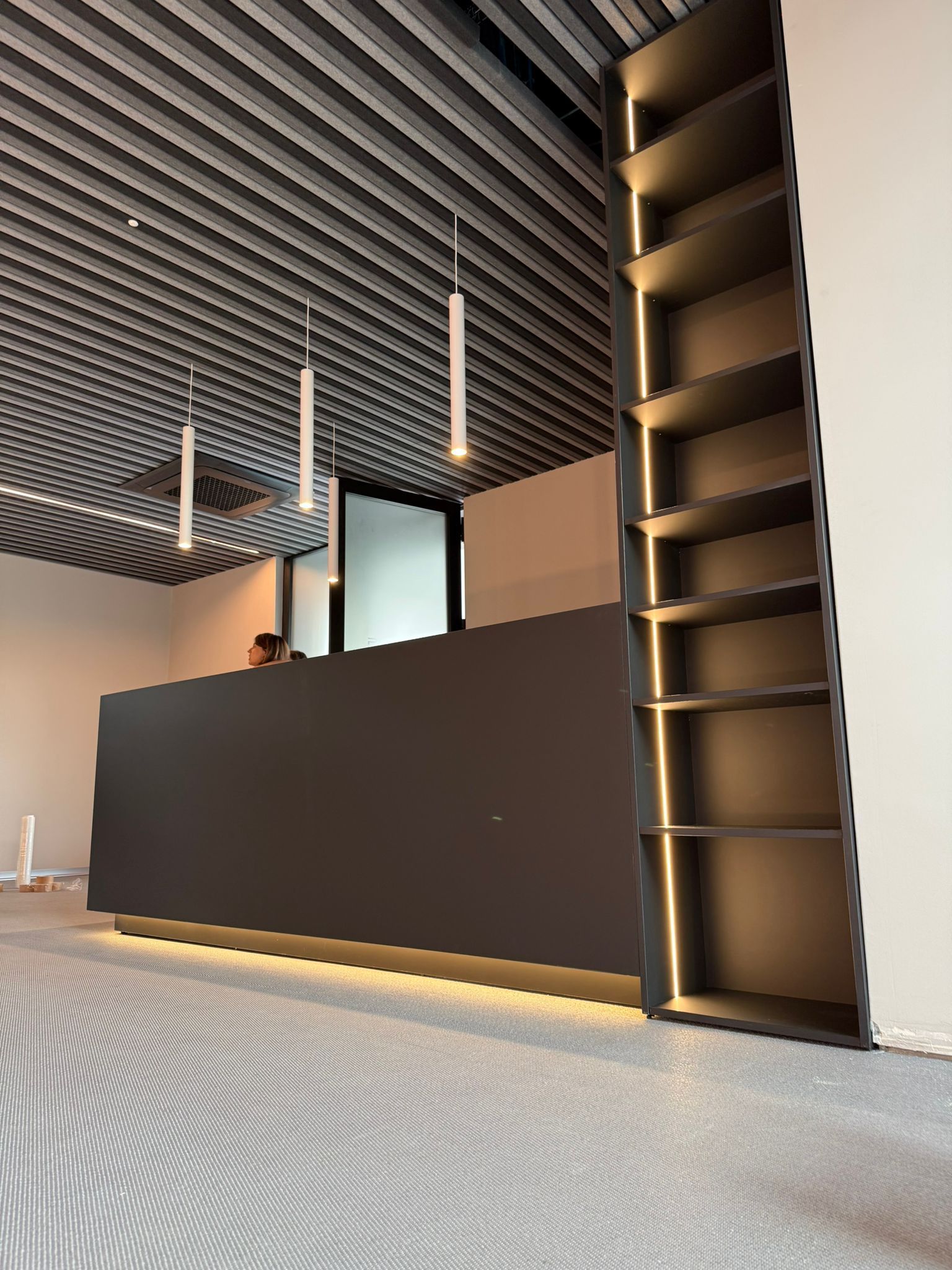 Un elegante bancone reception scuro in un ufficio moderno con libreria integrata retroilluminata, lampade a sospensione e soffitto con finitura a rilievo.