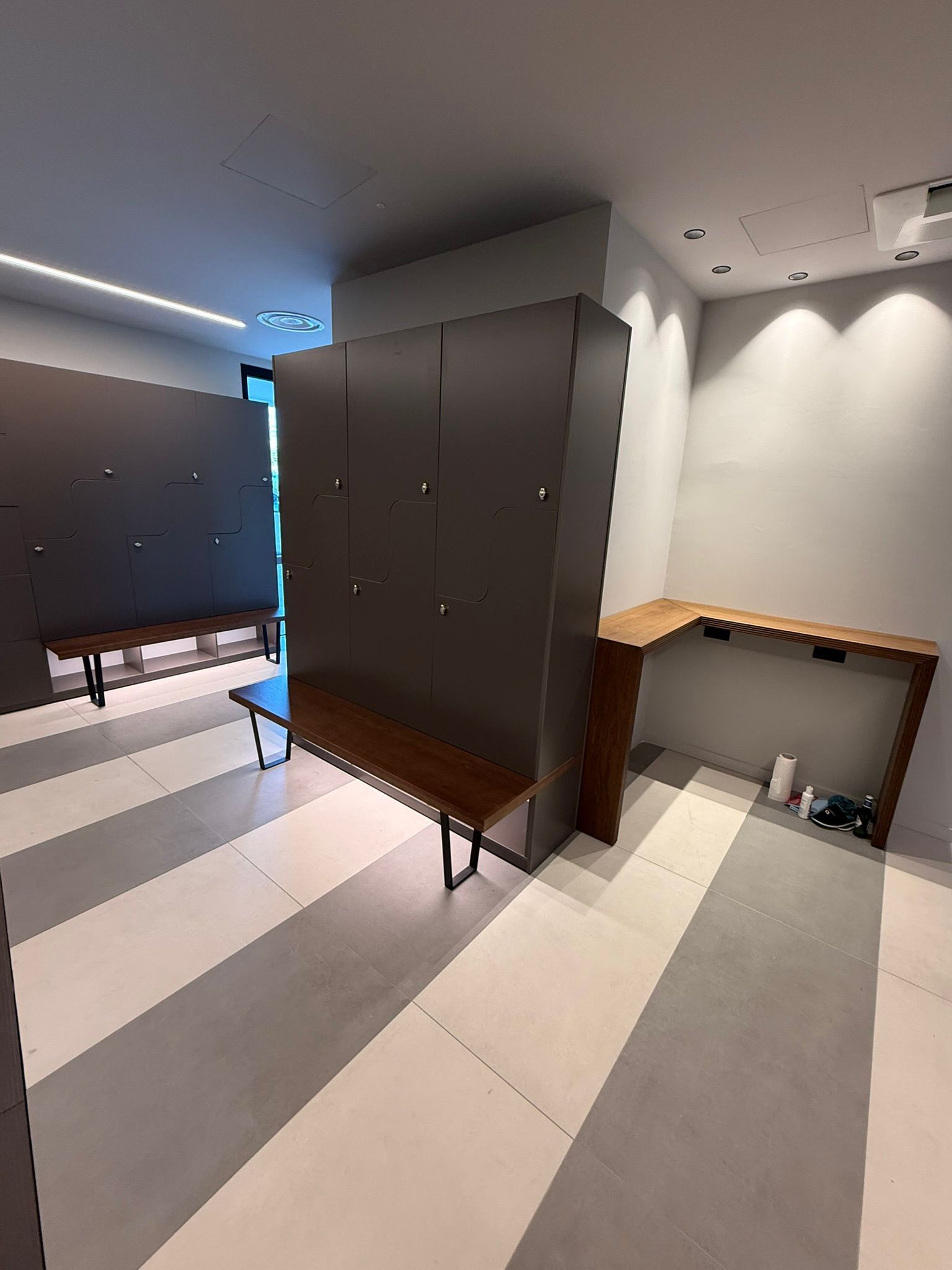 Uno spogliatoio moderno con armadietti grigio scuro, panche in legno e pavimento piastrellato a righe grigie chiare e scure.