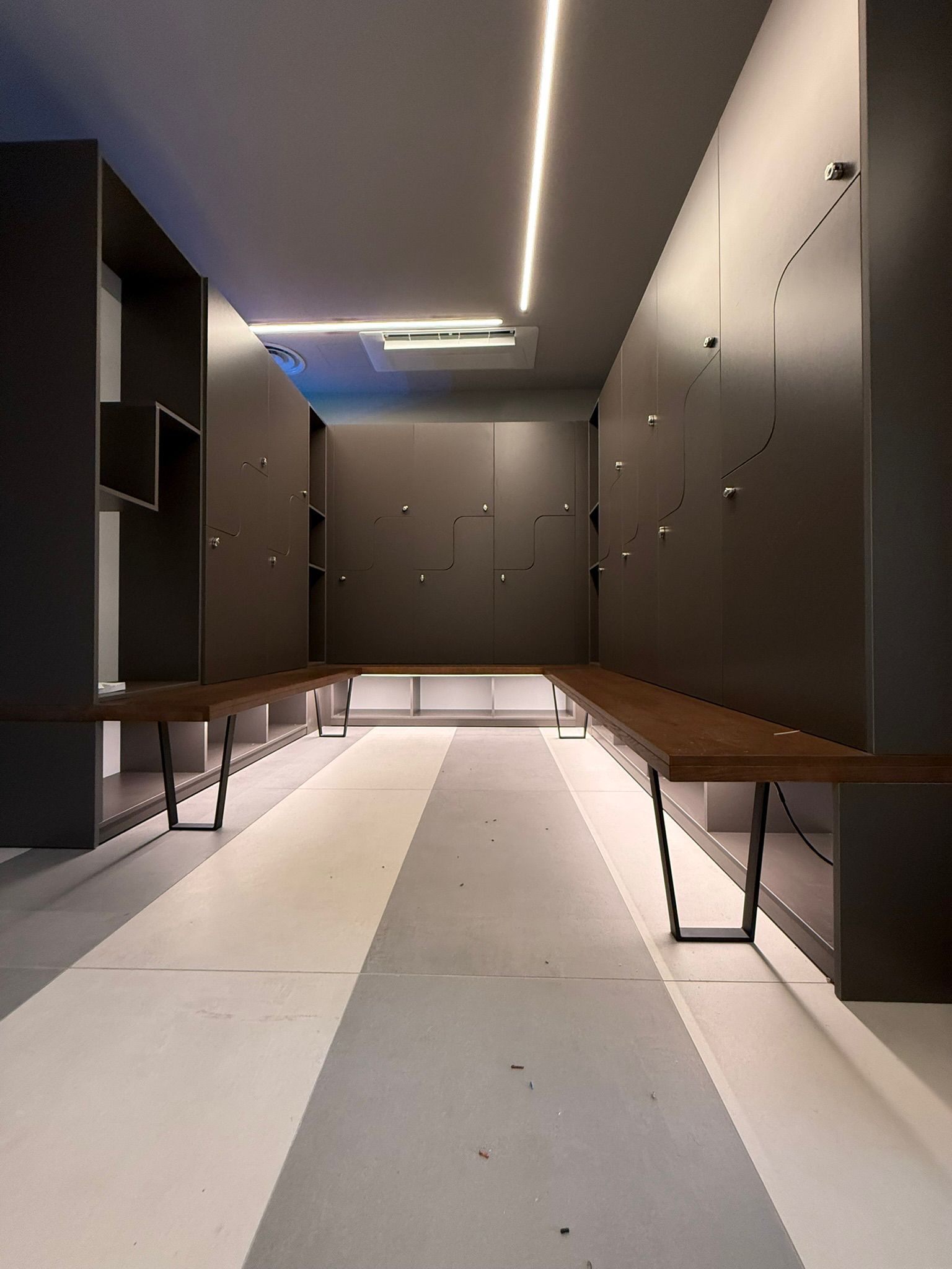 Spogliatoio moderno con armadietti grigio scuro, panche in legno e illuminazione d'accento a tutta altezza in un ambiente minimalista.