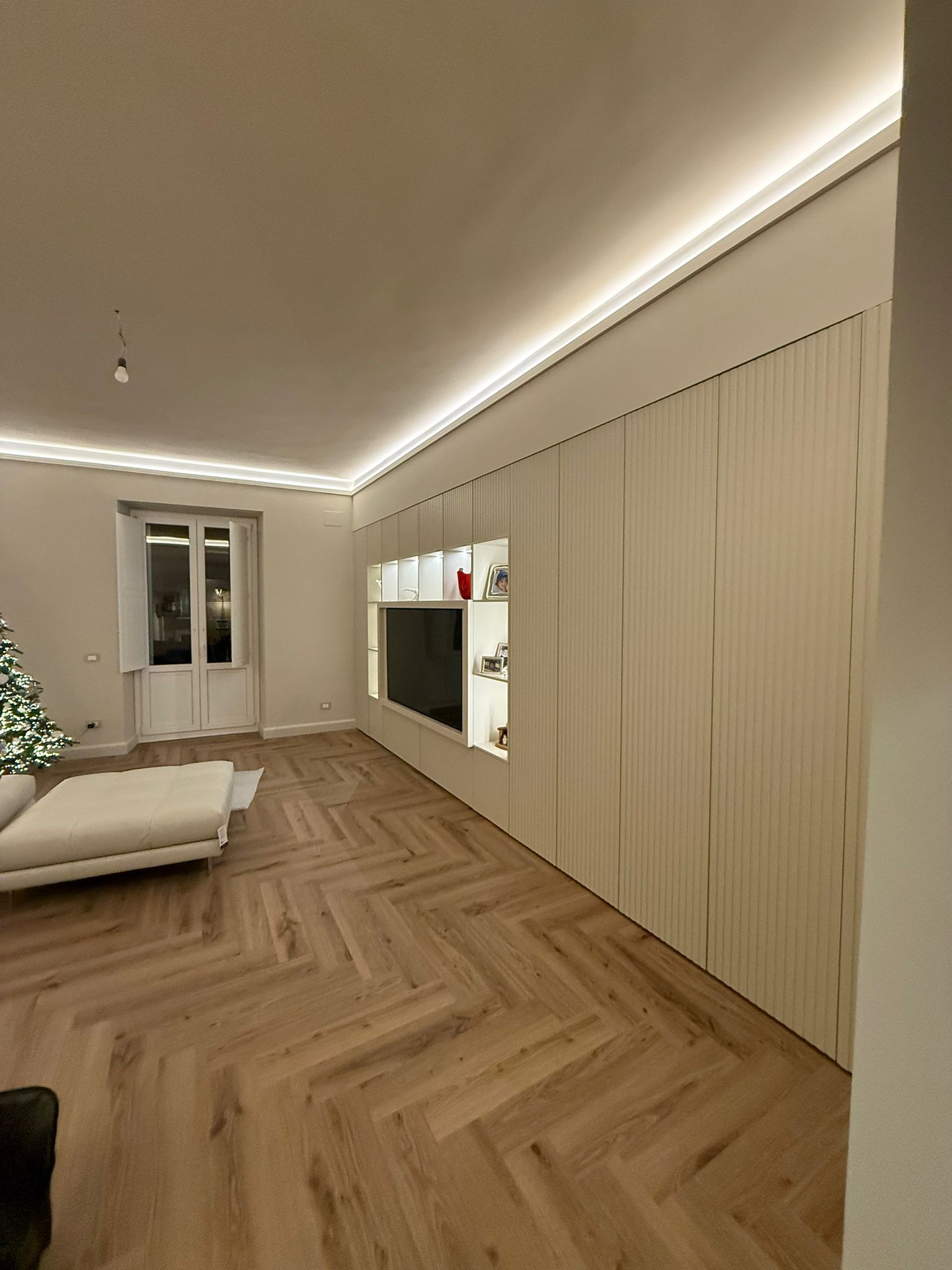 Soggiorno moderno con pavimento in legno a spina di pesce, grande mobile a muro in legno chiaro con ripiani incassati e illuminazione a LED.