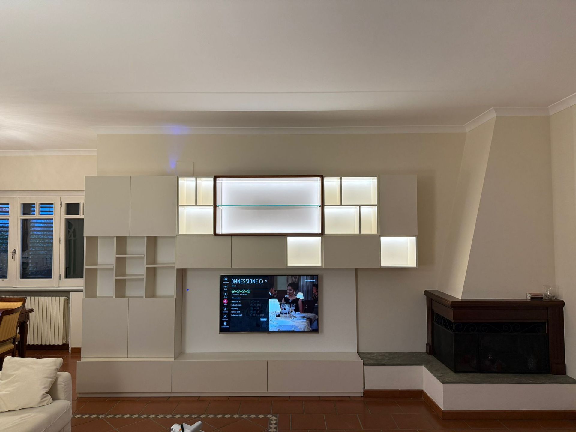 Una moderna parete attrezzata modulare color crema con illuminazione integrata e televisore a parete, posizionata accanto a un camino in pietra.