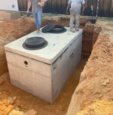Workers stand beside a buried concrete septic tank in a dirt excavation site.