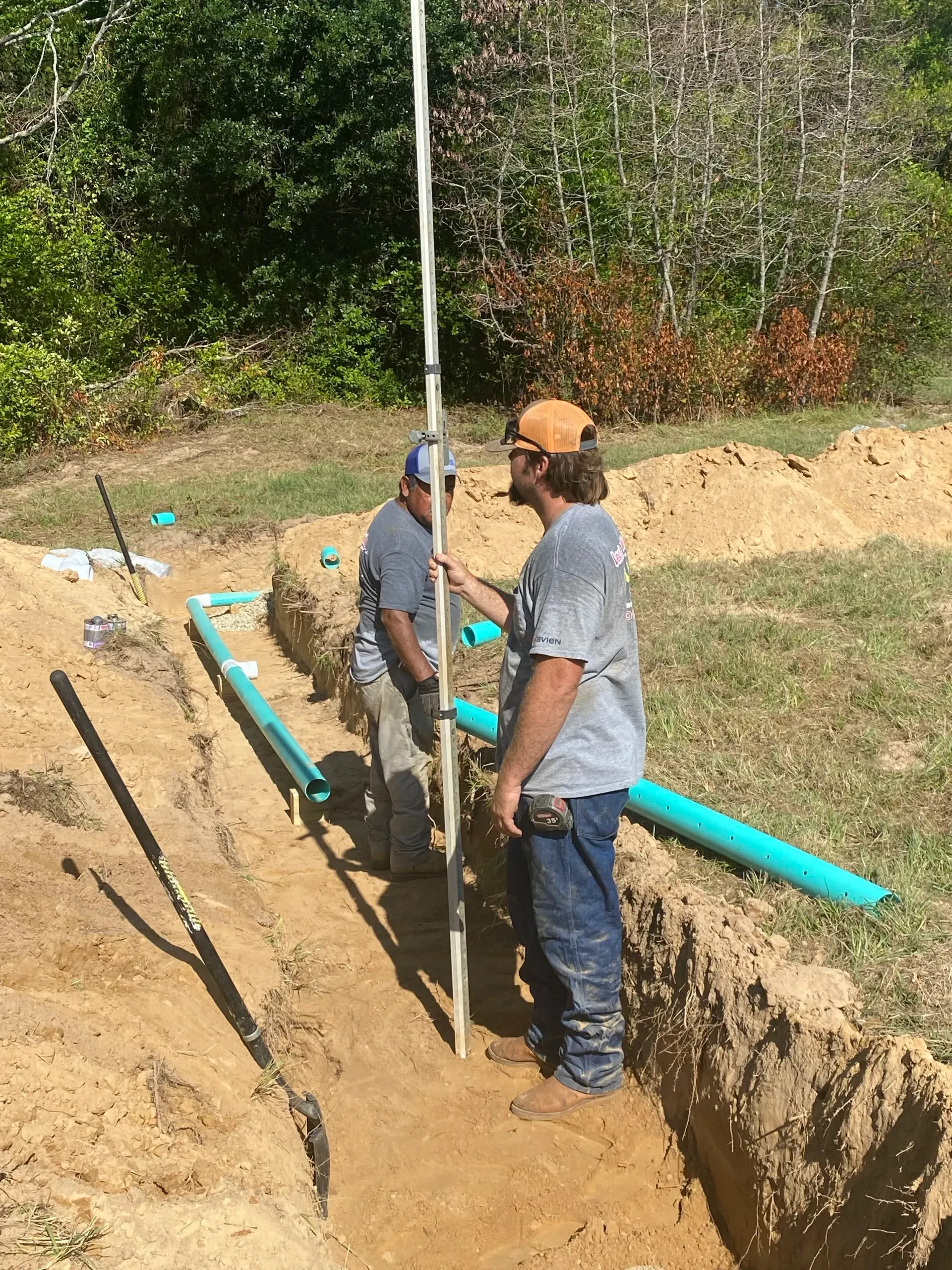 Two workers installing blue pipe in a trench outdoors next to a dirt path.