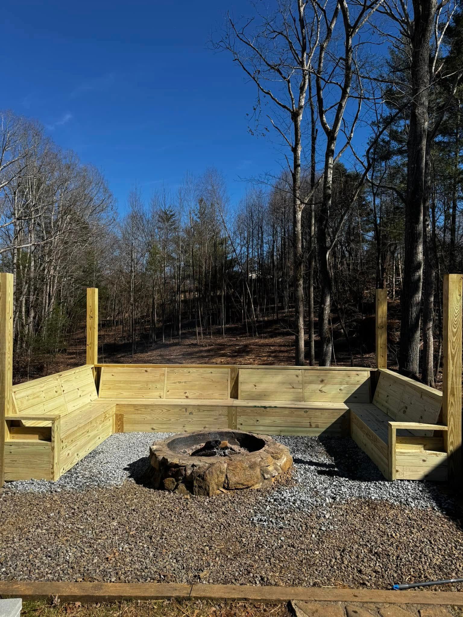Wooden fire pit seating area with gravel base, surrounded by trees under a clear blue sky.