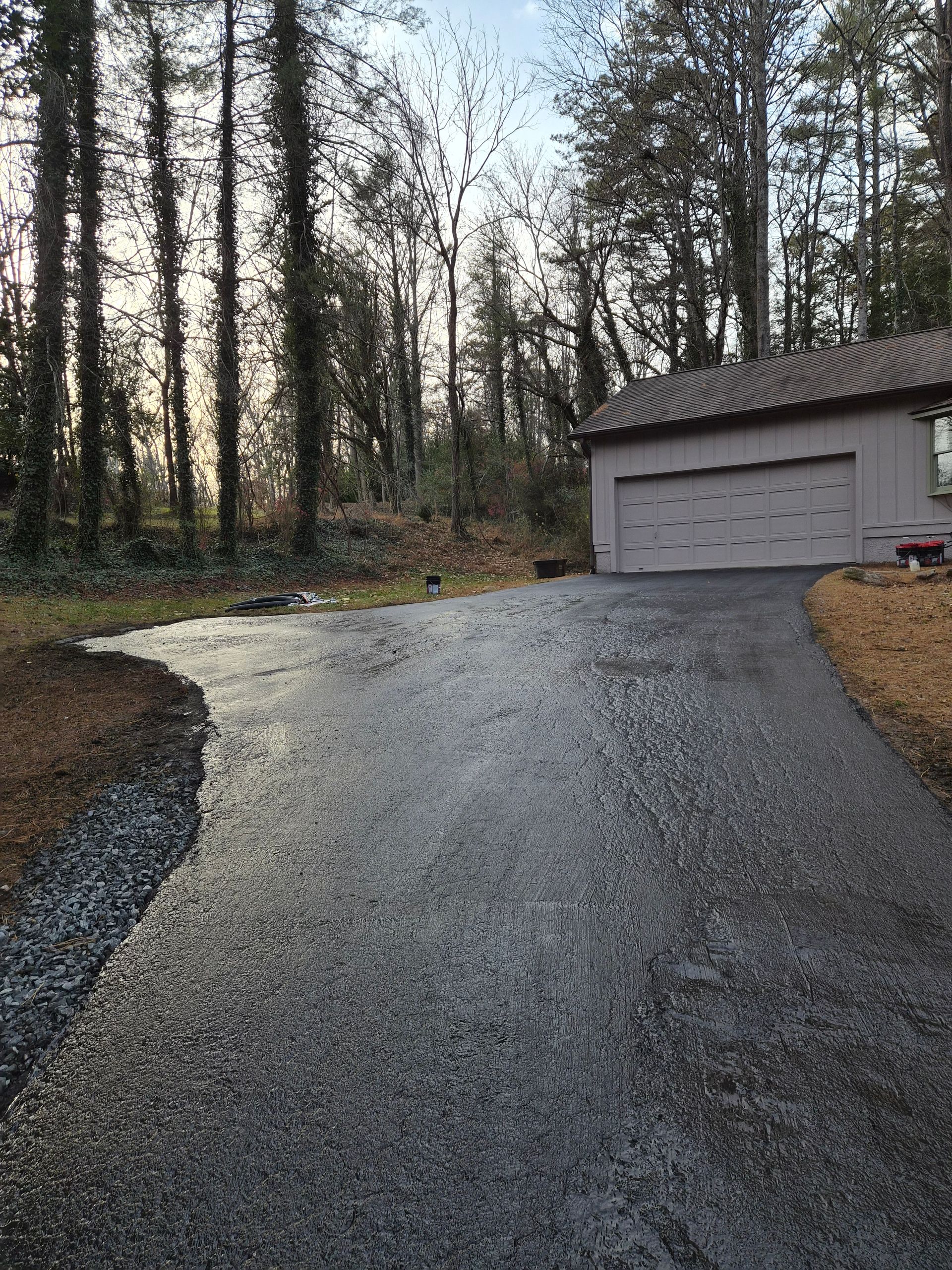 Asphalt driveway leading to a garage. Trees line the side and backdrop under a cloudy sky.