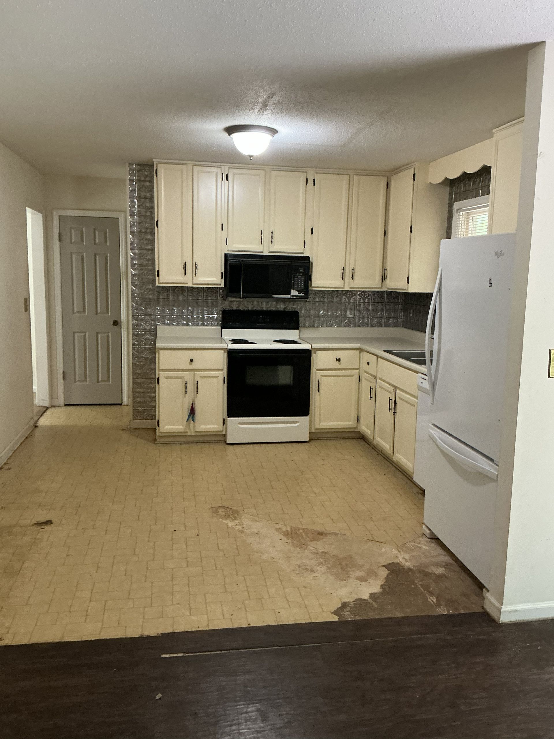 Kitchen with light cabinets, white appliances, and tiled backsplash, has stained flooring and a door in the background.