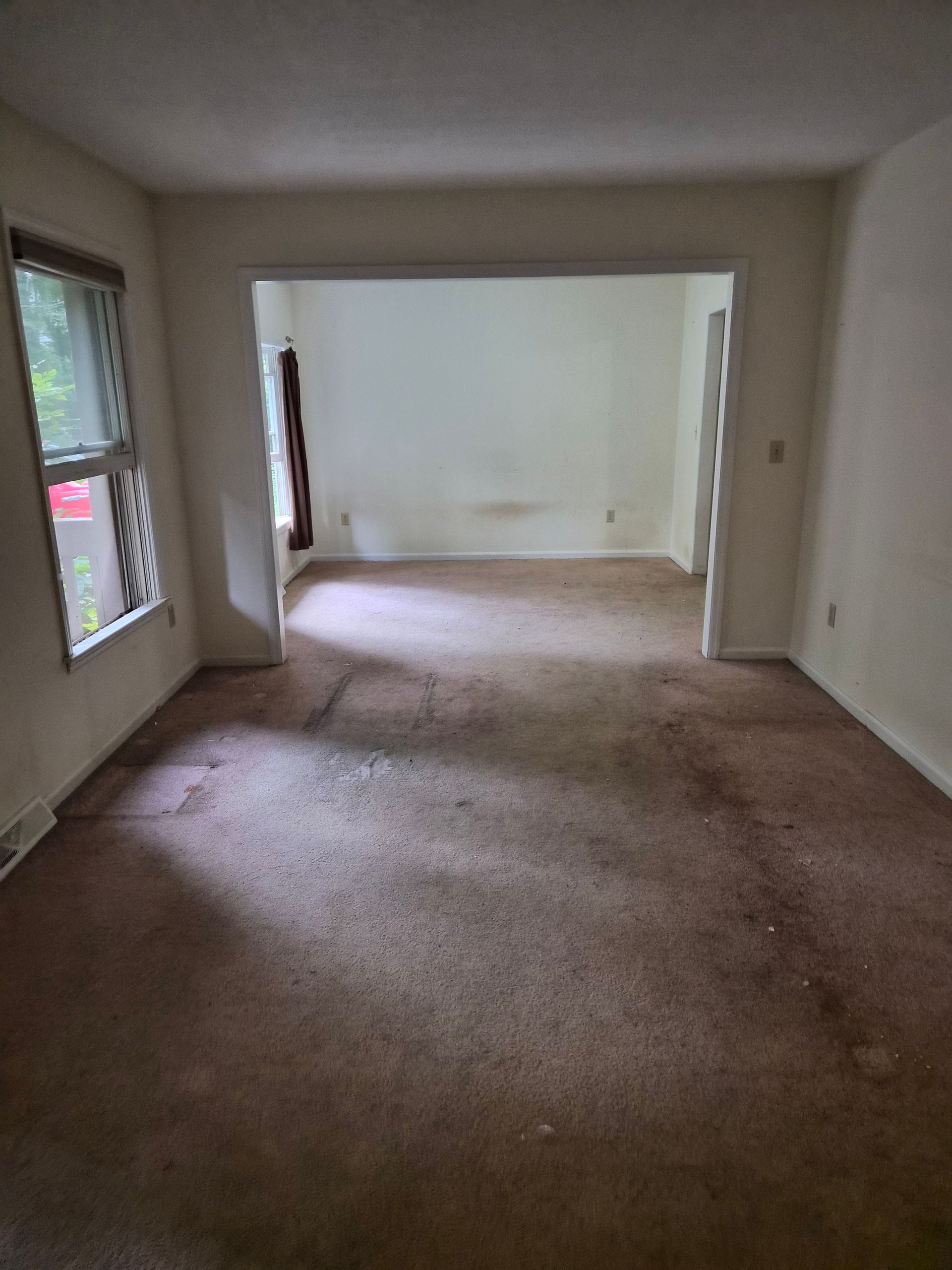 Empty room with stained brown carpet and white walls. Opens into a second room and a window.