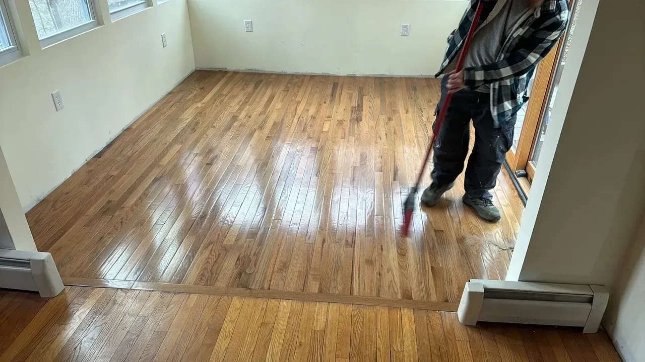 Person applying sealant to a hardwood floor in a room with windows and baseboard heaters.