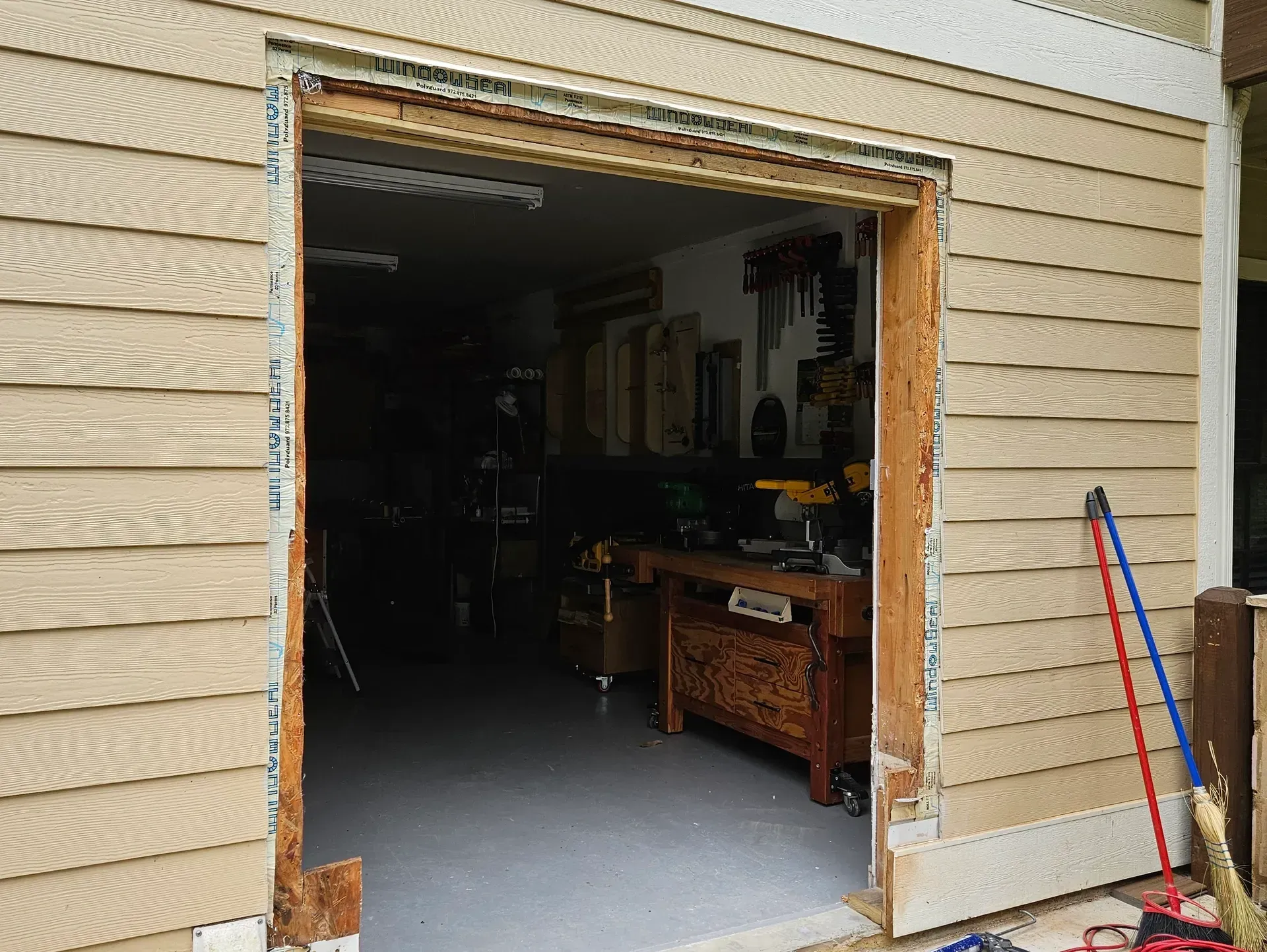 Open garage doorway, unpainted wood frame. Exterior siding is beige. Tools visible inside.
