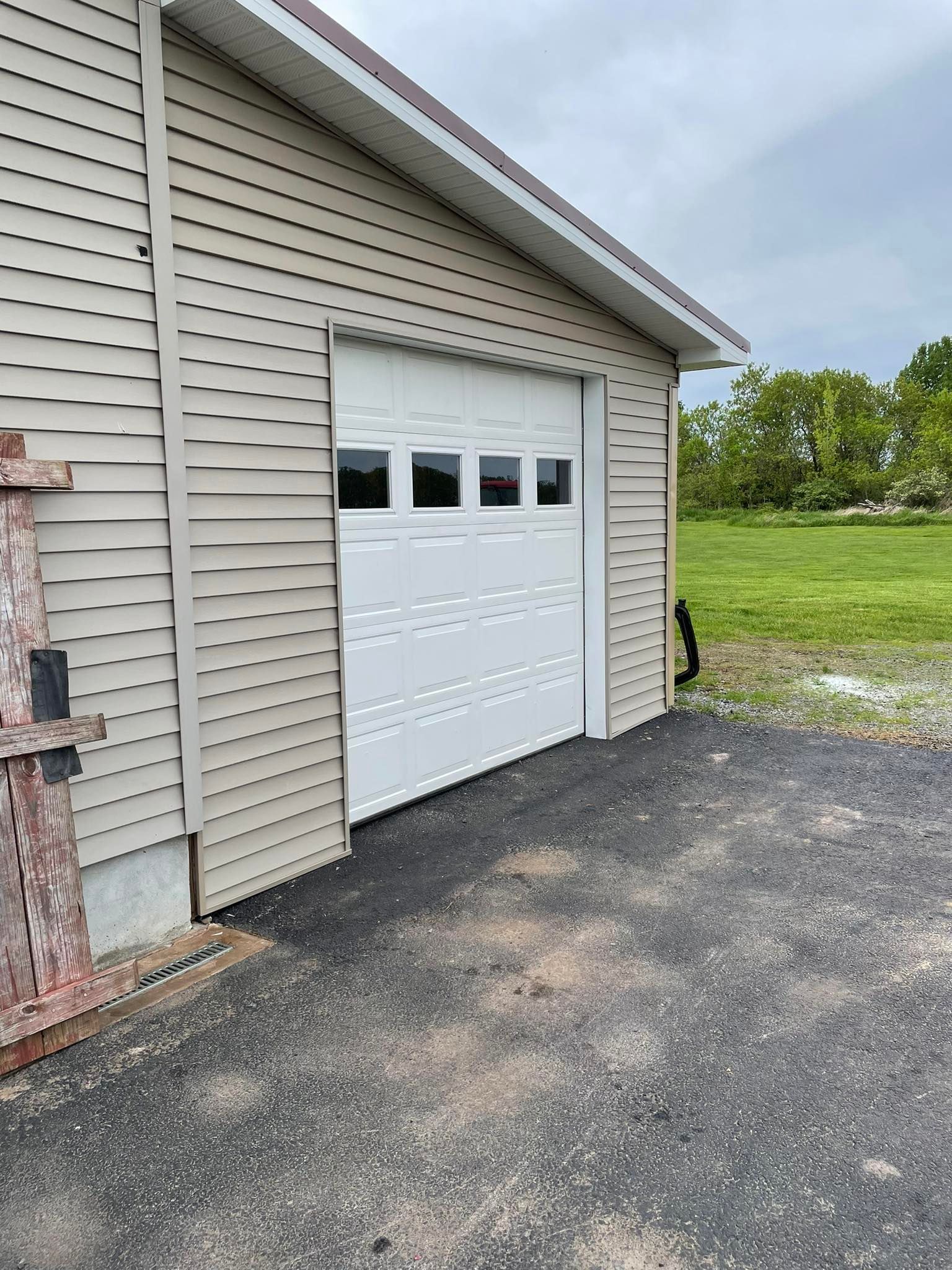 Garage with white door and tan siding on asphalt driveway, surrounded by grass and trees.