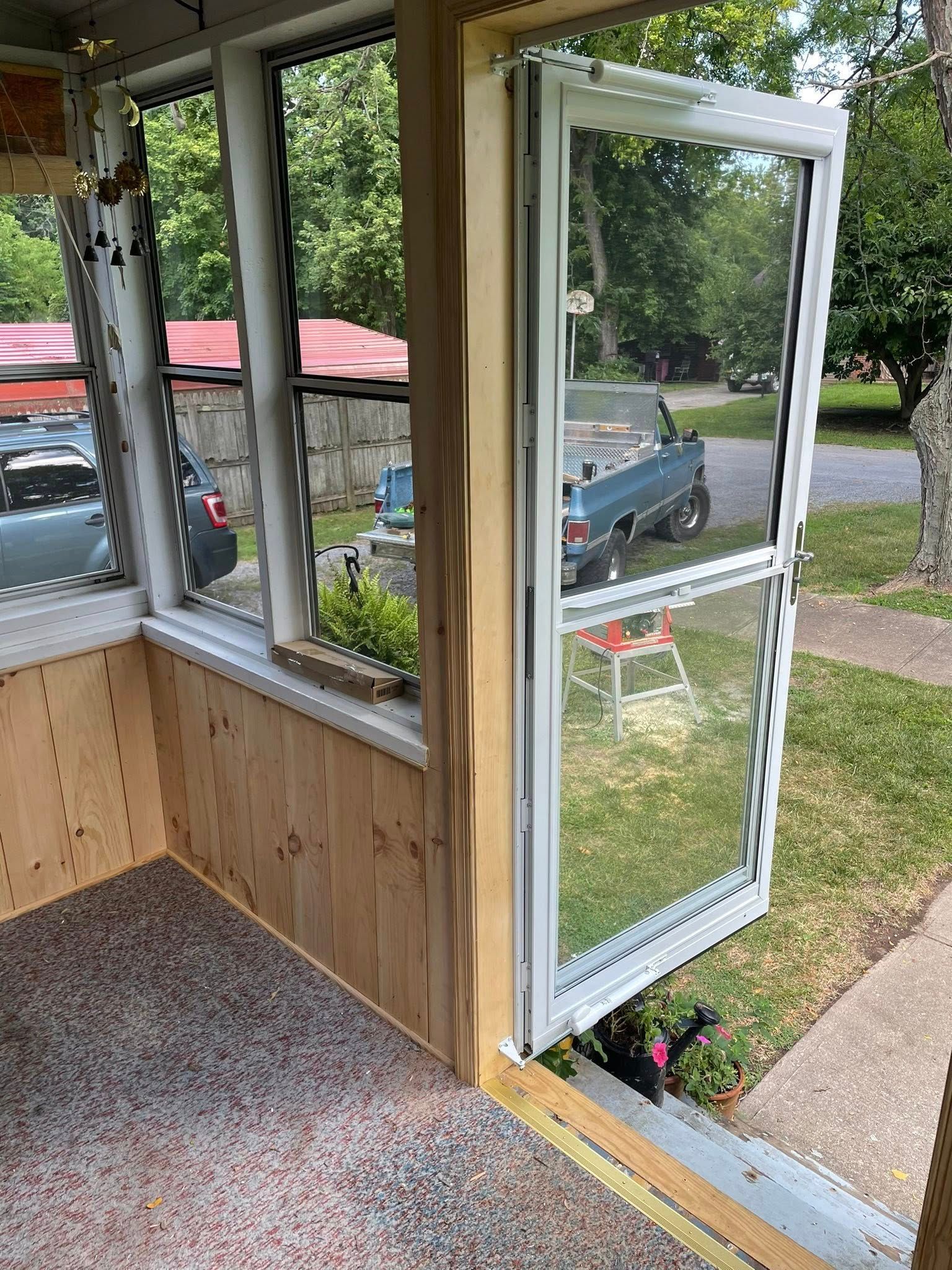 A porch with a screen door open to a yard, three windows, and wood paneling.
