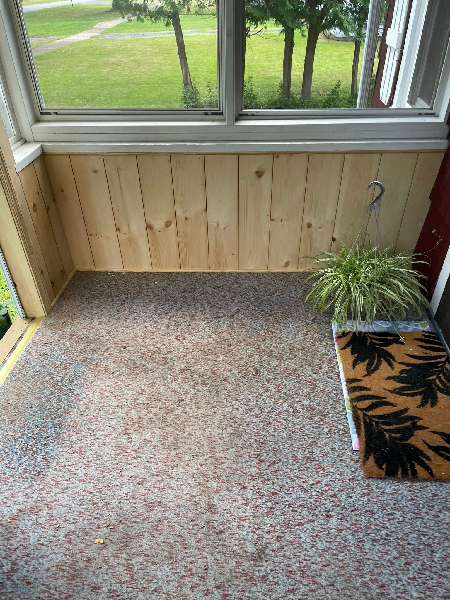 Enclosed porch with wood paneling, speckled floor, window view of trees, doormat, and potted plant.
