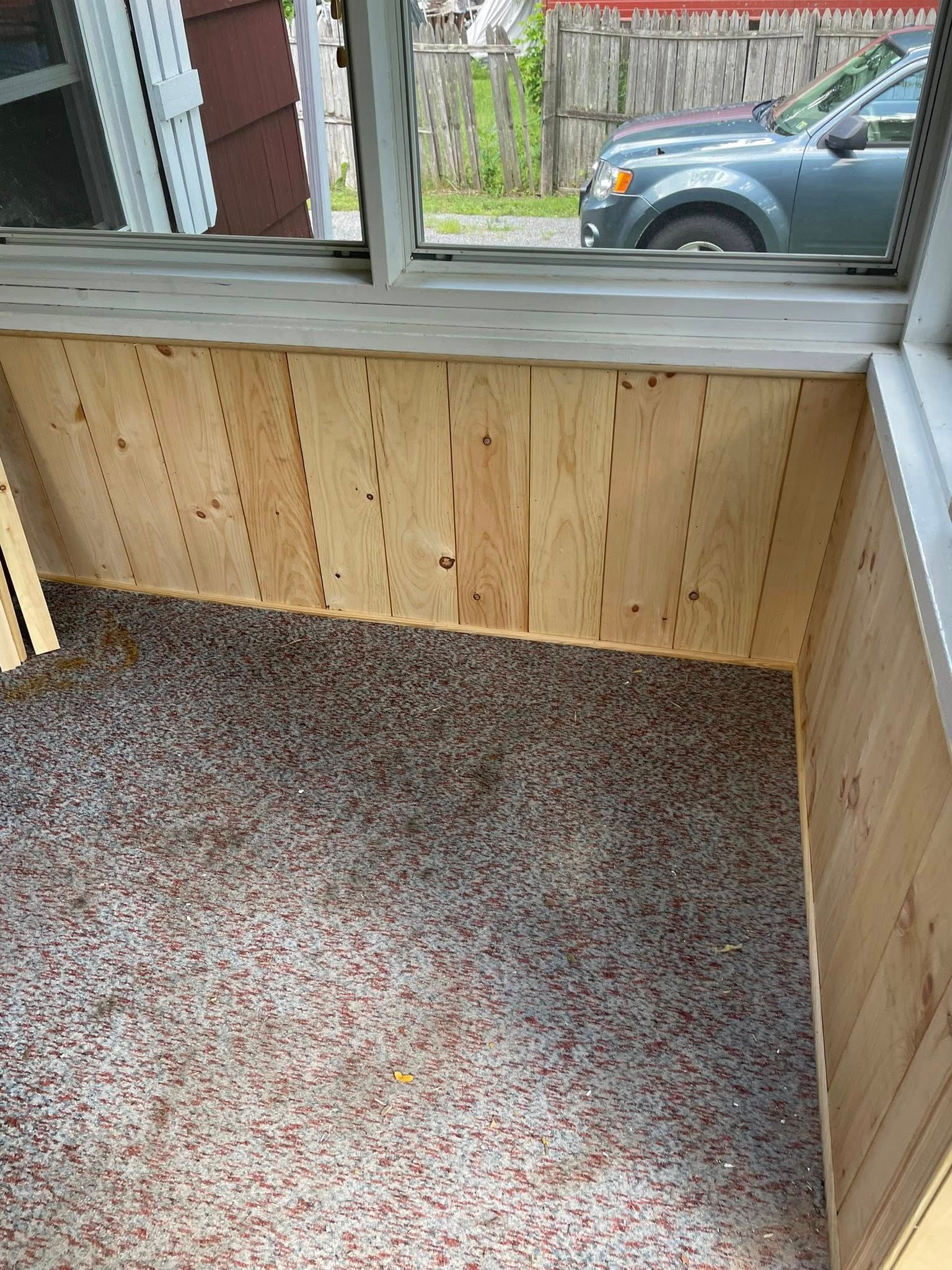 Interior view of a room with wood paneling and a speckled floor, looking out a window to a car and fence.
