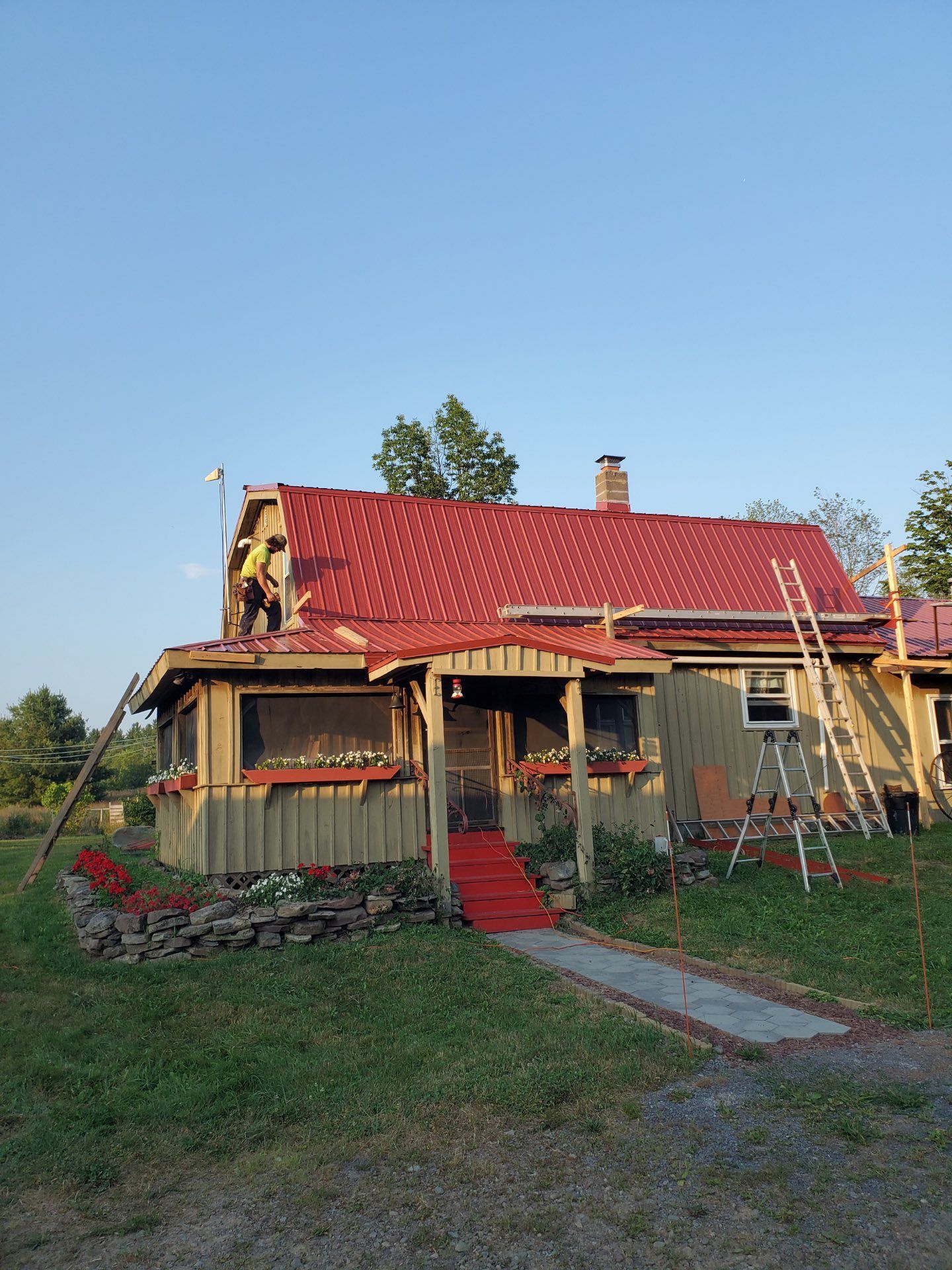 A red roof being installed on a weathered house with a porch, ladders, and landscaping on a sunny day.
