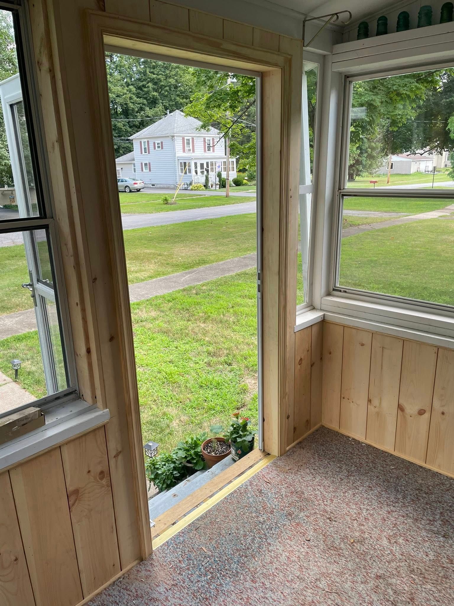 Sunroom interior with wooden trim and paneling, overlooking a grassy yard and a distant house.