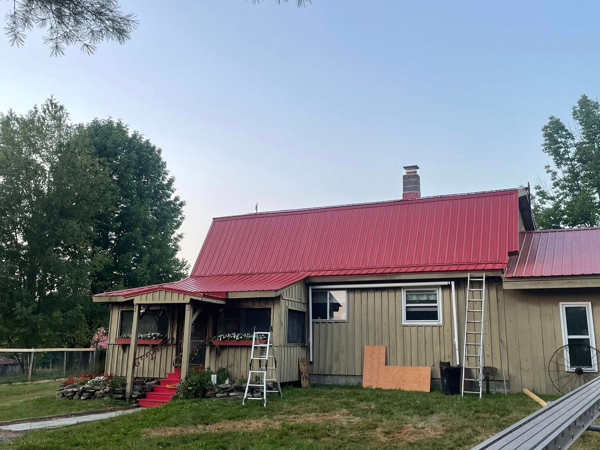 A one-story house with a red metal roof. A ladder stands near the side; outdoor setting.