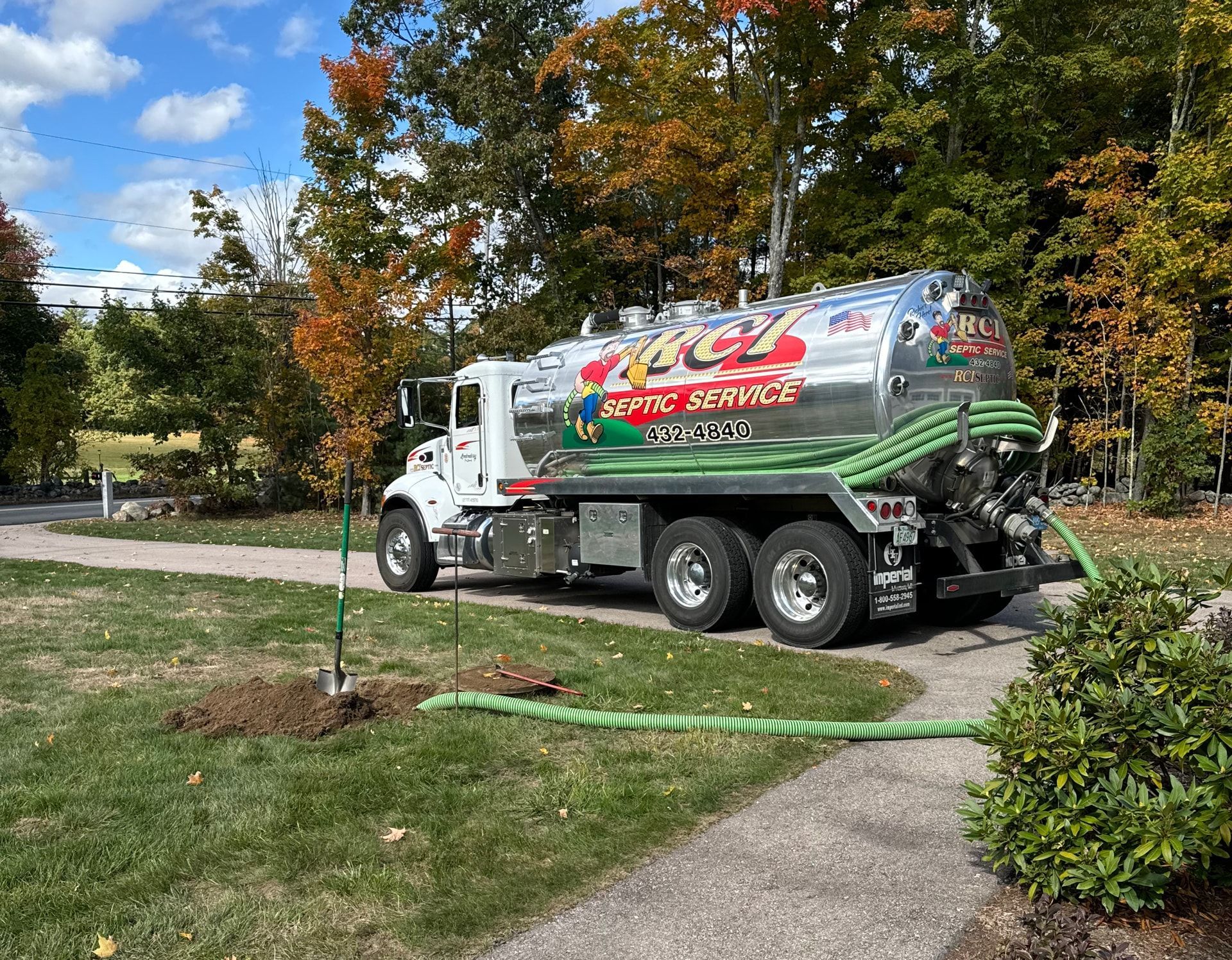 A white septic service truck parked on a paved path next to grass and bushes, with a green hose extending from the truck.