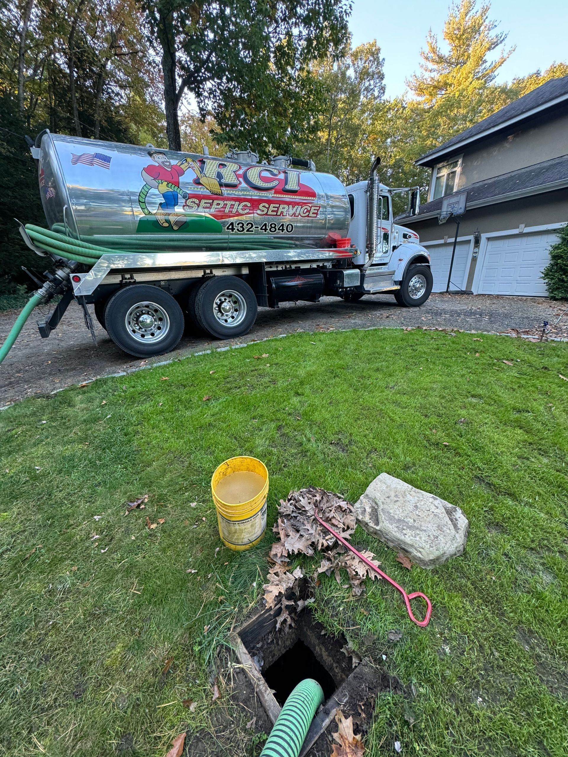 Septic tank truck parked on a lawn, with a hose connected to an open tank access. A yellow bucket and debris sit nearby.