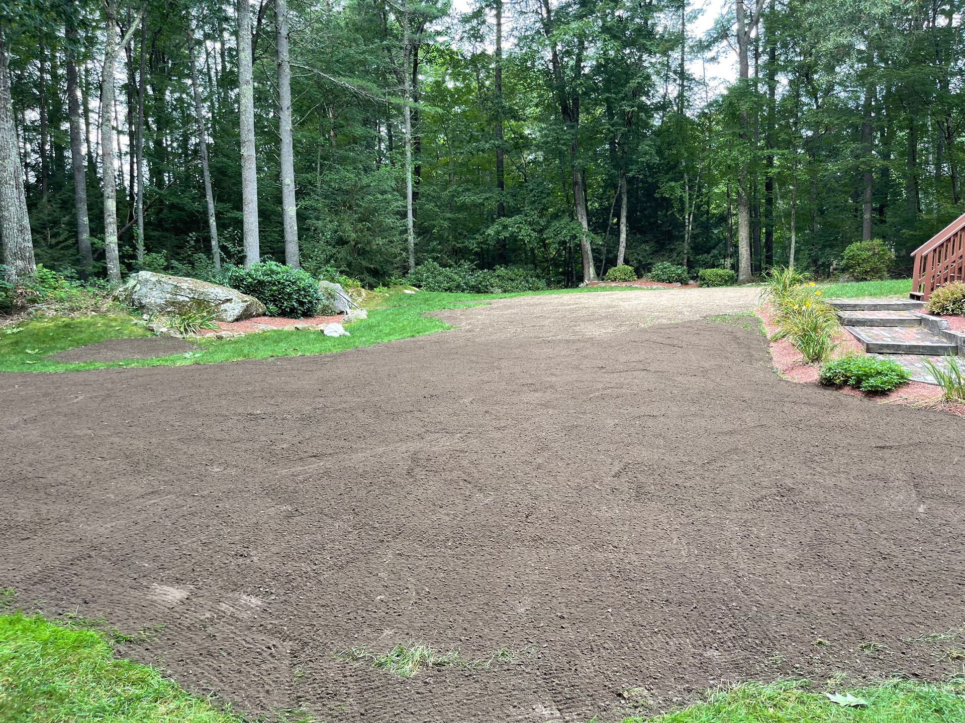 Freshly graded dirt driveway in a wooded yard with stone edging and a house deck on the right