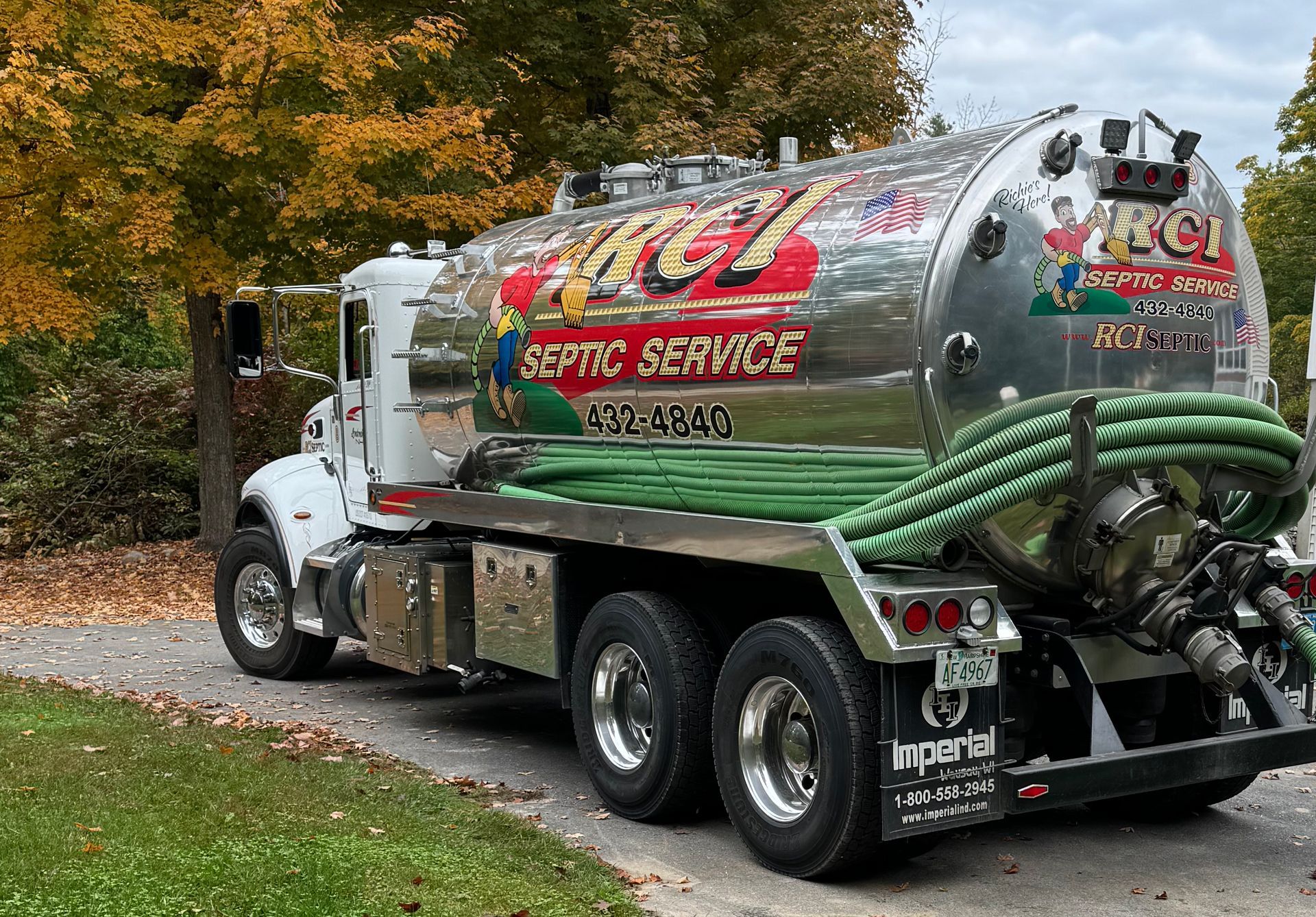 A shiny septic service truck parked on a driveway with fall foliage in the background. The truck is white and silver, with green hoses.