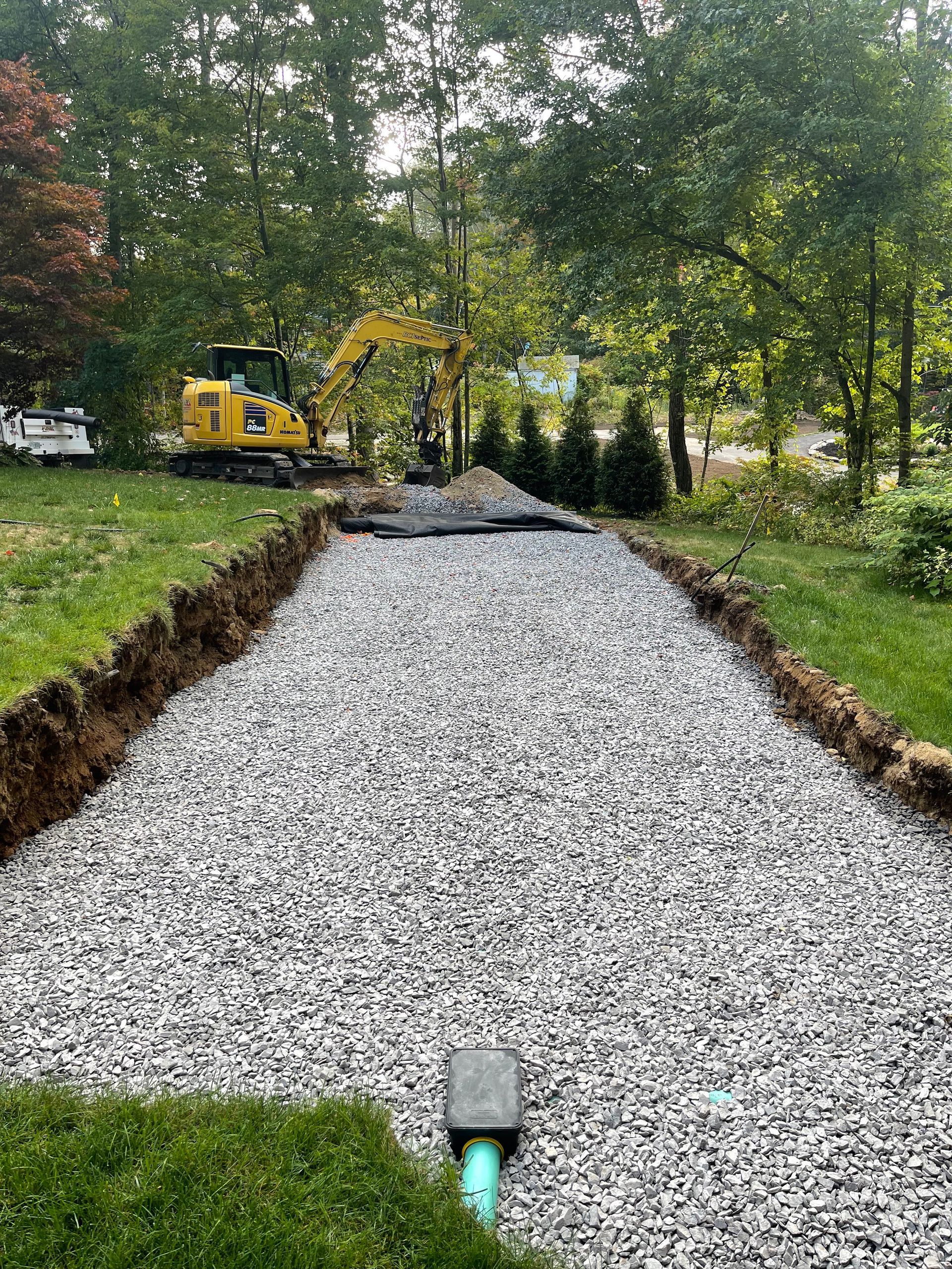 Gravel-filled trench with green pipe, likely a leach field. Excavator in background, surrounded by grass and trees.