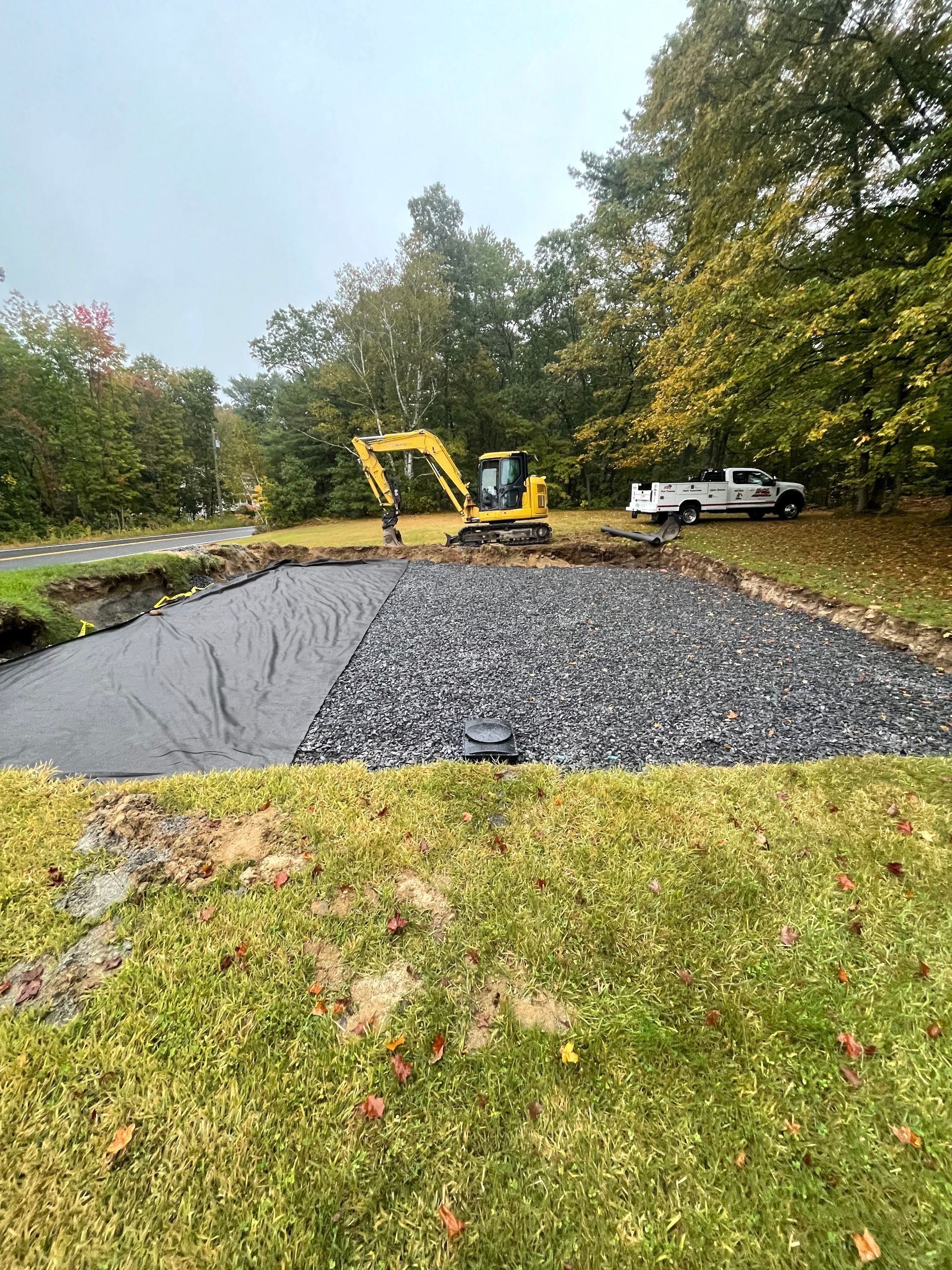 Excavator and truck constructing a gravel-covered area, likely for a driveway or parking, on a green lawn.