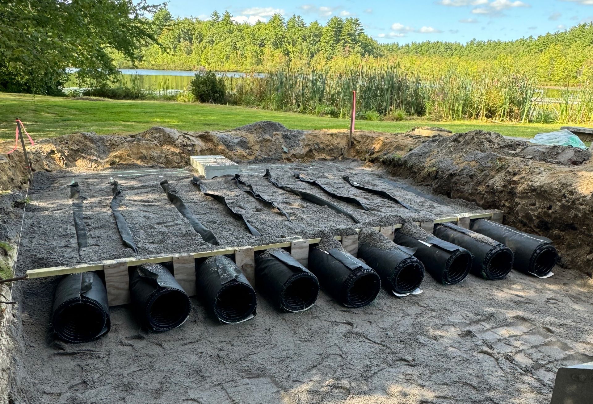 Black pipes installed in a dirt trench, supported by wood, near a grassy area and water.