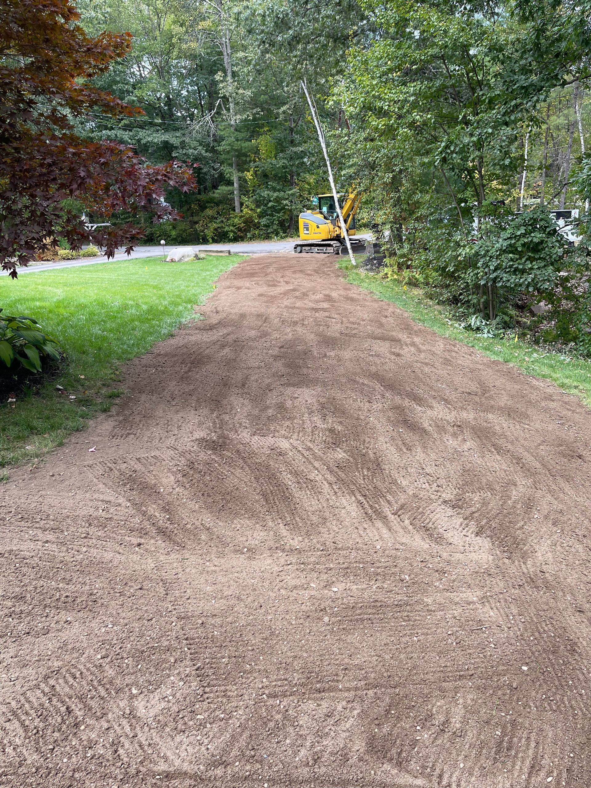 Gravel driveway being prepared with a small excavator in the background, surrounded by trees and grass.