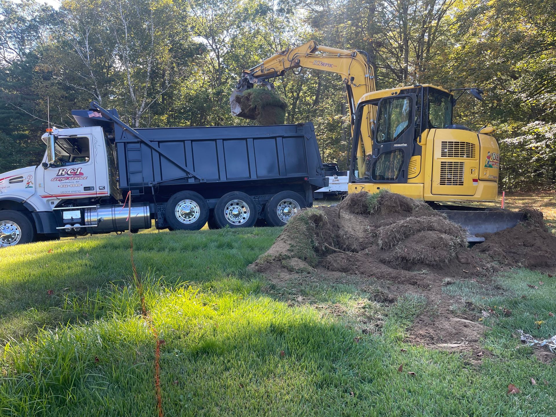 Yellow excavator loading dirt into a black dump truck in a grassy area near trees.