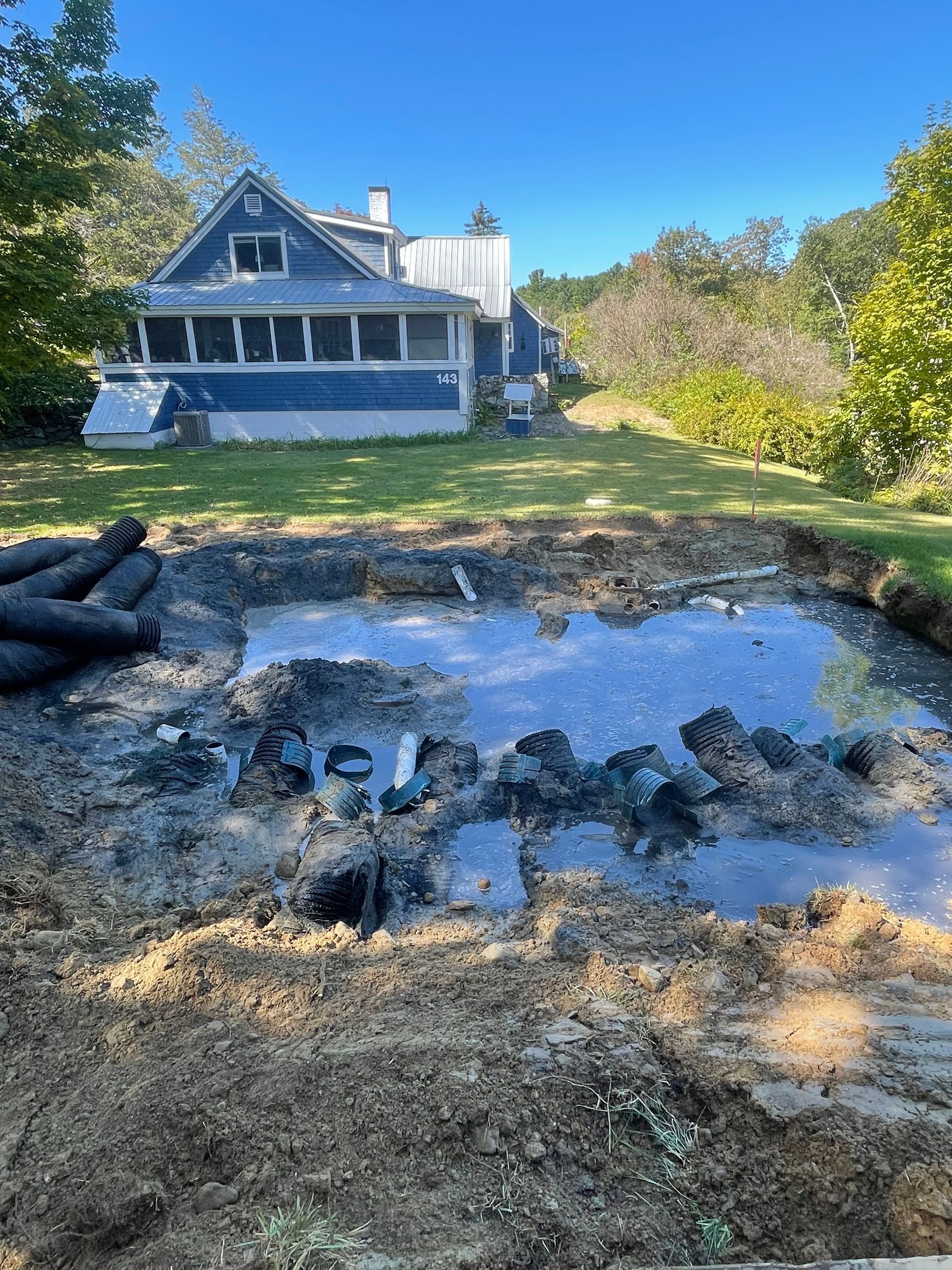 A dug-out hole filled with water in a yard, a blue house in the background on a sunny day.
