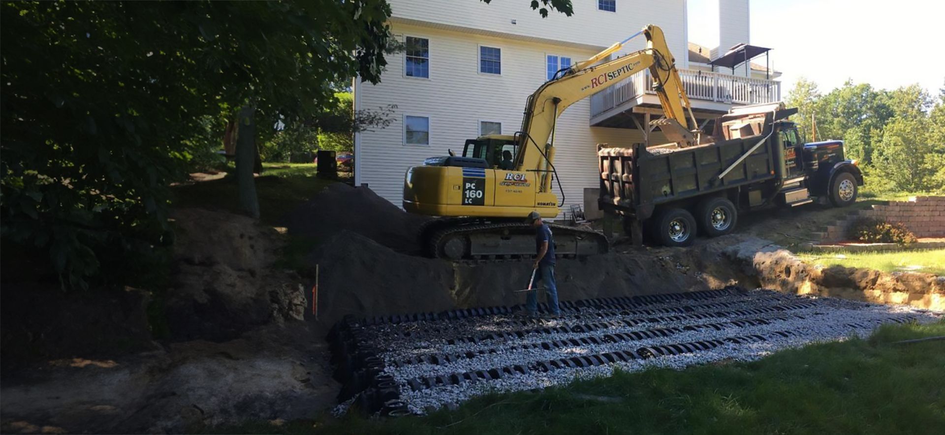 a yellow excavator is loading dirt into a dump truck in front of a house  that is getting a septic system installed