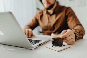 A man is sitting at a desk using a laptop computer while holding a cup of coffee.