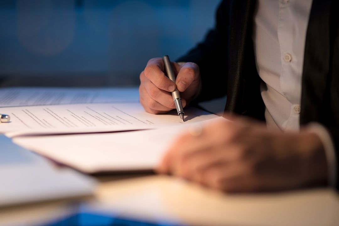 A professional in a suit signs a document with a silver pen at a desk in a dimly lit office.