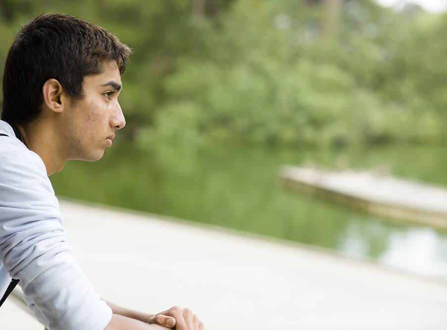 A person looks pensively over a railing toward a pond and a small wooden dock in a park.