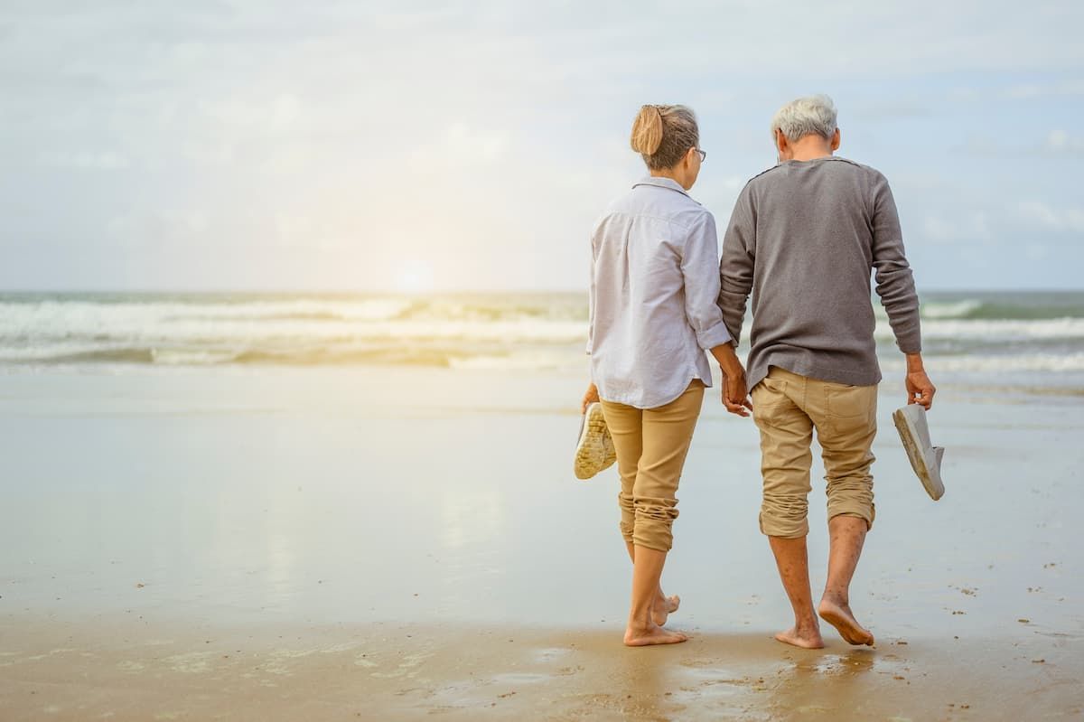 An older couple walks barefoot along a sandy beach, holding shoes and walking toward the ocean under a soft, hazy light.
