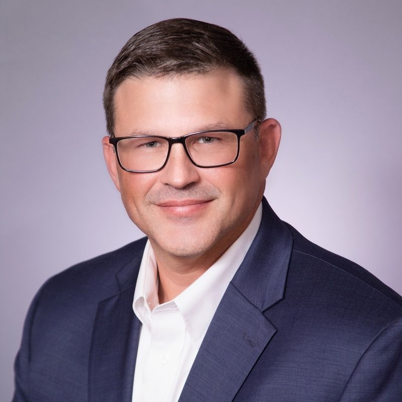 Man in glasses and suit smiling, headshot against a gray background.