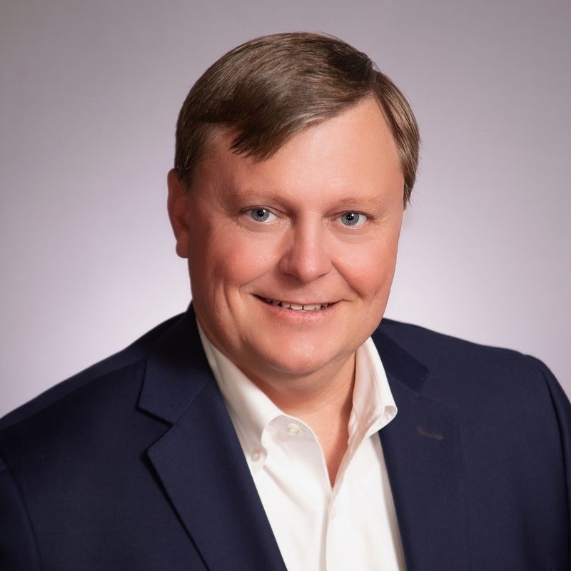 Man in a navy blazer and white shirt smiles at the camera, against a gray backdrop.