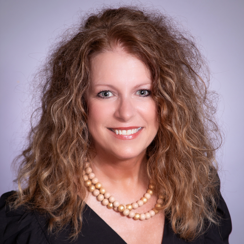 Woman with long, curly brown hair, wearing a layered beaded necklace and a black top, smiling.