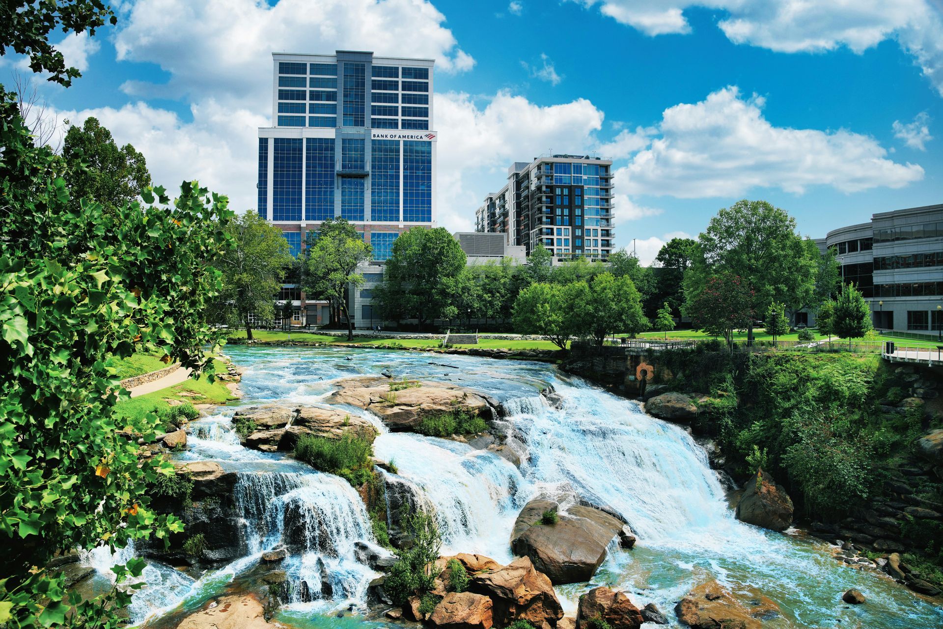 Waterfalls cascade in front of modern buildings on a sunny day with blue sky and green trees.