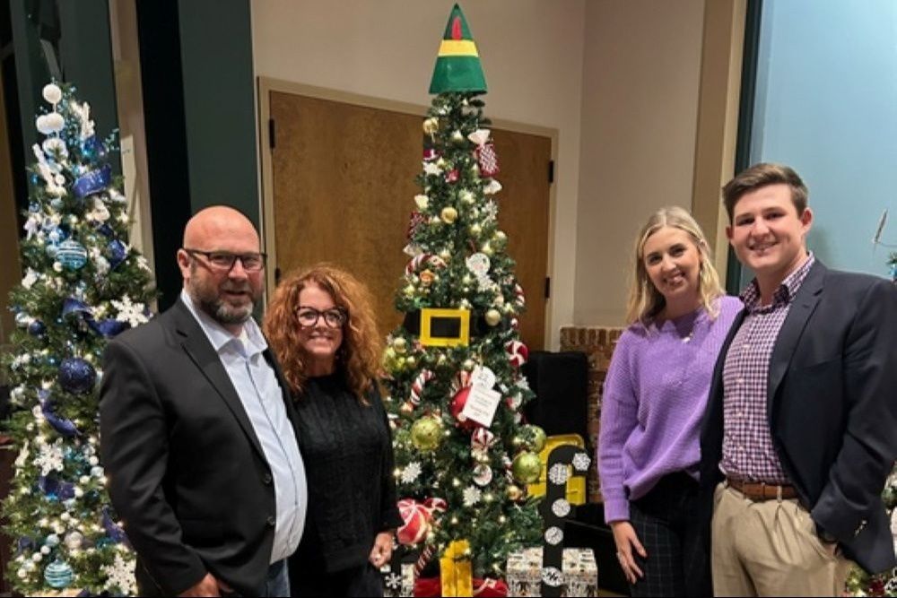 Four people pose by Christmas trees, one with an elf hat topper.