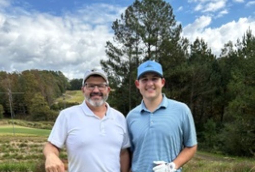 Two men on a golf course. One wears a white shirt and hat, the other a blue shirt and cap, holding a golf club.