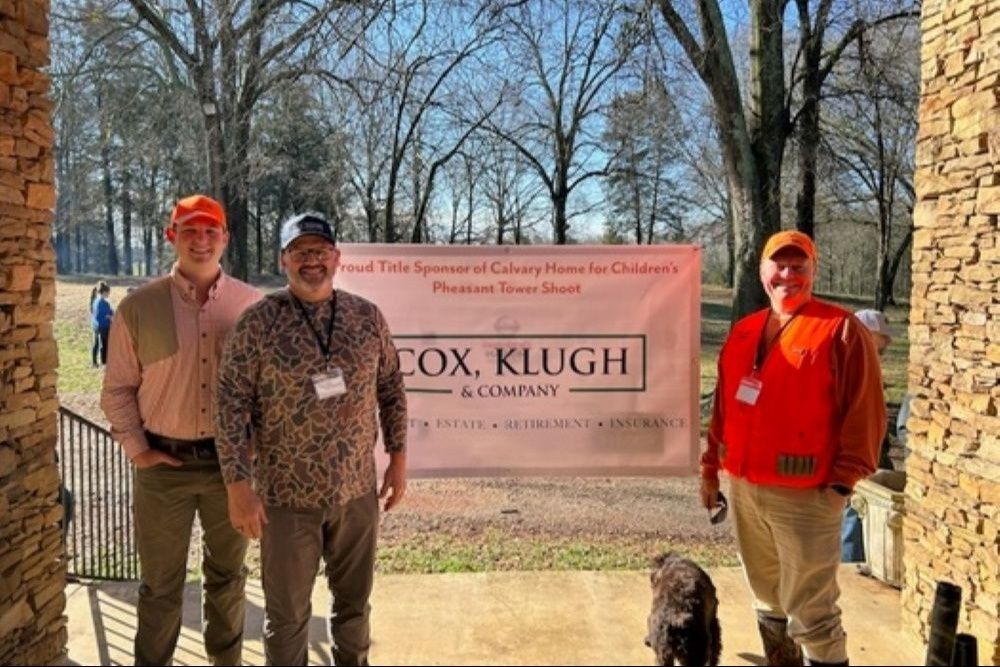 Three men pose outdoors with a banner for a company. One wears an orange vest, one a leopard print shirt.