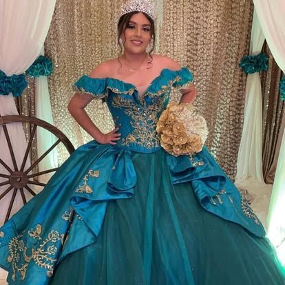 A woman in a teal and gold ball gown is standing in front of a wagon wheel.